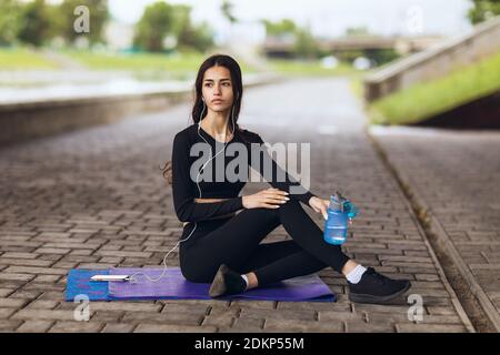 A young woman sits on a gymnastic mat on the waterfront and rests after doing sports. Stockfoto
