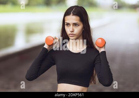 Beautiful young woman performs exercises with dumbbells in the street. Healthy lifestyle concept. Stock Photo