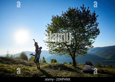 Rückansicht von zwei glücklichen Reisenden in der Nähe von Camping und großen Baum in den Bergen am sonnigen Morgen ruhen. Guy hält Mädchen in den Händen. Tourismus Abenteuer aktiven Lebensstil Konzept Stockfoto