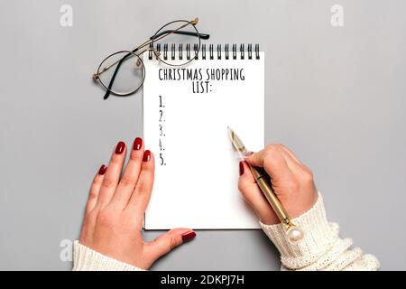 Weibliche Hand hält Stift, schreiben Weihnachtseinkaufsliste, Geschenkideen auf weißem Notizblock auf grauem Hintergrund Draufsicht Flat Lay Weihnachtskarte Frau macht Chr. Stockfoto