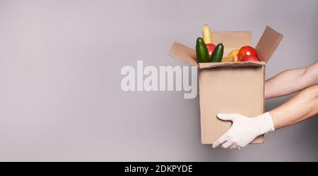 Hände von weißen Kurier in Gummihandschuhen halten einen Karton mit Lebensmitteln, schnelle und sichere Lieferung von Lebensmitteln. Stockfoto