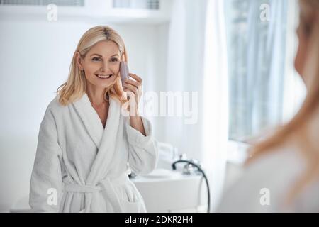 Glückliche lächelnde Frau in weißem Bademantel Blick auf den Spiegel Im Badezimmer Stockfoto