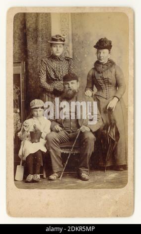 Original charaktervolles viktorianisches CDV (Carte de Visite) Erinnerungsfoto einer Arbeiterfamilie mit Schwiegermutter im Urlaub im Badeort Skegness, Lincolnshire, England, um 1888. Die Mutter ist attraktiv. Vater ist bärtig. Das Kind, das einen Eimer und Spaten hält, trägt eine merkwürdige Mischung aus Mädchenkleid und Knabenmütze und ist angesichts der viktorianischen Gewohnheit, Jungen in Mädchenkleidung zu kleiden, wahrscheinlich ein Junge. Er sieht nicht sehr glücklich aus und lässt sein Foto offenbar unter Druck machen.