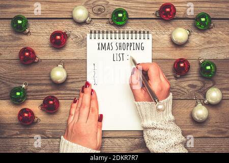 Weibliche Hand hält Stift, schreiben Weihnachtseinkaufsliste, Geschenkideen auf weißem Notizblock auf Holzhintergrund Draufsicht Flat Lay Weihnachtskarte Frau machen Stockfoto