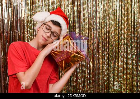 Junge mit einem Geschenk für das neue Jahr auf einem goldenen Hintergrund. Junge Teenager mit Brille und in einem Weihnachtsmann Hut mit . Weihnachtsdekoration.