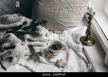 Gemütliches Winterfrühstück Stillleben Szene. Tasse heißen Kaffee mit Lebkuchen in der Nähe des Fensters. Weihnachtskonzept. Kissen, Strickpullover und Tanne Stockfoto