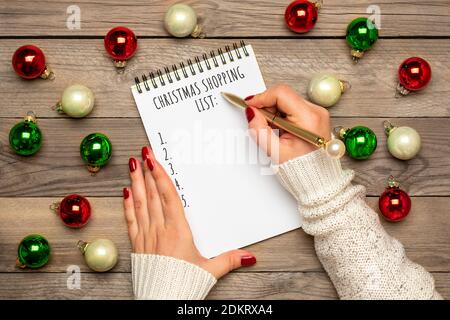 Weibliche Hand hält Stift, schreiben Weihnachtseinkaufsliste, Geschenkideen auf weißem Notizblock auf Holzhintergrund Draufsicht Flat Lay Weihnachtskarte Frau machen Stockfoto