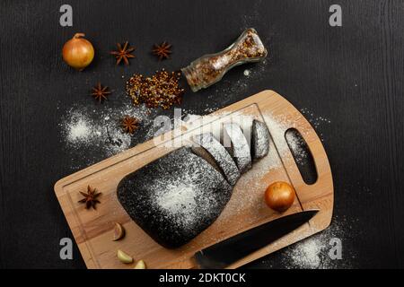 Frisches schwarzes Roggenbrot auf einem Schneidebrett mit Gewürzen Mit Mehl auf einem schwarzen Holztisch bestreut Stockfoto
