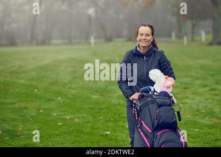 Fröhlich freundlich lächelnde weibliche Golfspielerin mit Tasche und Keulen an Ein üppig grüner Golfplatz an einem kalten nebligen Winter Tag im aktiven Lifestyle-Konzept mit Cop Stockfoto