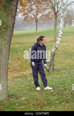 Frau Golfspielerin spielt eine Runde Golf, die zwischen Bäumen steht An einem kalten Wintertag mit Keule in der Hand starren Raus über den Fairway Stockfoto