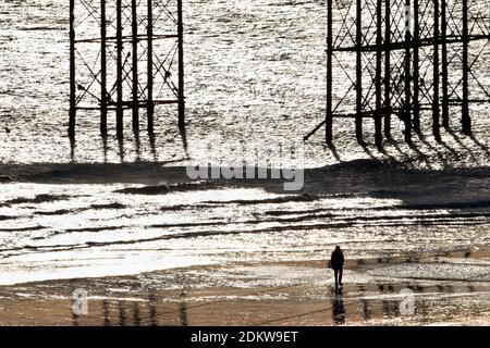Silhouette eines einsamen Mannes, der am Strand in der Nähe der Säulen des Palace Pier bei Ebbe in Brighton, Sussex, England, Großbritannien spaziert Stockfoto