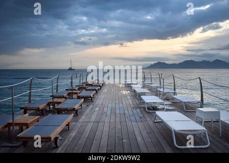 Sonnenliegen auf leerem Holzponton unter bewölktem Himmel im Herbst in Cannes, Cote d'Azur, Frankreich Stockfoto