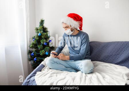 Junger Kaukasier mit Gesichtsmaske und Weihnachtsmann-Hut auf der Couch sitzend, am Telefon des weihnachtsbaums chatschend Stockfoto