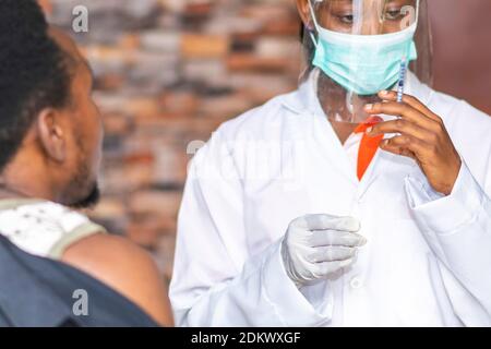 afrikanisches medizinisches Personal, das eine Impfspritze hält und sich bereit macht, sie einem jungen schwarzen Mann zu verabreichen Stockfoto