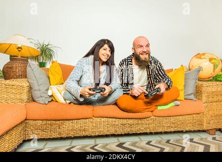 happy couple of a hipster bald man and caucasian brunette woman holding two console joypad sitting comfortable on a couch for gaming evening.fun. Stockfoto