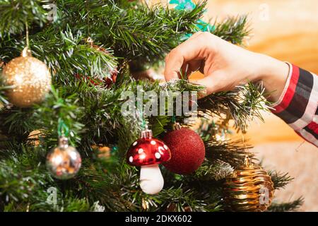 Nahaufnahme einer Frau in einem karierten Hemd hängt eine schöne glänzende rote Kugel auf einem Weihnachtsbaum auf einem hölzernen Wandhintergrund. Weihnachten und Neujahr zu Hause vor Stockfoto