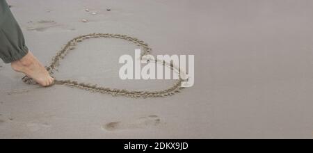 Eine unkenntliche Frau zieht am 14. Februar mit dem Fuß ein Herz auf den Sand. Valentinstag 2021. Stockfoto