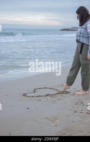 Vertikales konzeptionelles Bild. Erkennbare Frau zieht am 14. Februar mit dem Fuß ein Herz auf den Sand. Valentinstag 2021. Stockfoto