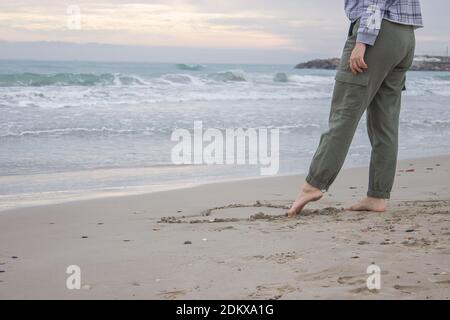 Horizontales konzeptionelles Bild mit Ozeanhintergrund. Eine unkenntliche Frau zieht am 14. Februar mit dem Fuß ein Herz auf den Sand. Valentinstag Stockfoto