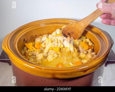 Nahaufnahme der Hand eines Mannes, der einen heißen Auflauf / Eintopf mit heißem Huhn und Gemüse, mit einem Holzspatel, in einem elektrischen langsamen Herd rührt. Stockfoto