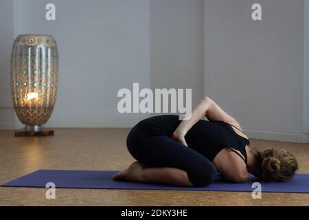 Yogi Frau praktiziert Kind Pose Variation im Studio mit Lampe durch weißen Hintergrund. Yoga-Studentin in Balasana mit einem Twist. Flexibilitätskonzept Stockfoto