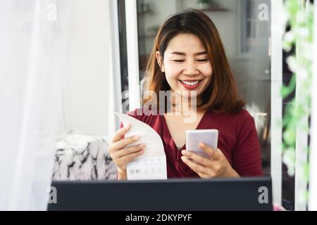 Glücklich asiatische Frau zahlen Strom Rechnung Hand halten Smartphone Code wird auf Papier gescannt Stockfoto