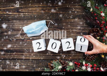 Frau Hand kippt Neujahr 2020 Holz Kalender Blöcke zu 2021. Medizinische Maske, Weihnachtsbaumlichter, Dekorationen, Kiefernzweige, rote Winterberrie Stockfoto