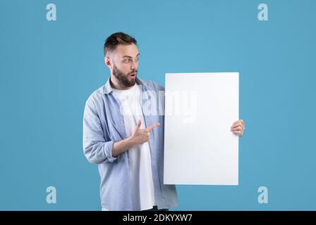 Überrascht Millennial Mann zeigt auf weiße leere Poster auf blauem Studio-Hintergrund, mockup für Ihre Anzeige-Design Stockfoto