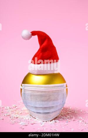 Goldene Weihnachtskugel mit blauer chirurgischer Gesichtsmaske und Weihnachtsmann Hut auf rosa Hintergrund. Stockfoto