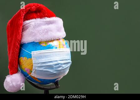 Erdkugel mit Weihnachtsmannhut und blauer chirurgischer Gesichtsmaske auf grünem Hintergrund. Das Konzept der Feier des neuen Jahres und Weihnachten während der COV Stockfoto