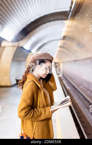 Junge Frau im Mantel und Baskenmütze liest Buch über U-Bahn Plattform Stockfoto