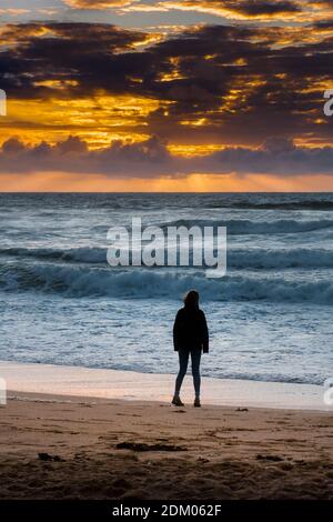 Eine Frau, die allein am Fistral Beach steht und von einem spektakulären Sonnenuntergang in Newquay in Cornwall umgeben ist. Stockfoto