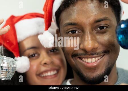 Nettes lächelndes lateinisches Paar mit weihnachtsmützen auf den Köpfen, die Weihnachtskugeln halten und die Kamera betrachten. Konzentriere dich auf das Gesicht des Jungen. Stockfoto