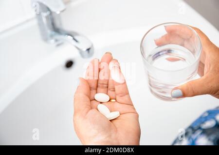 Female elderly hand with pills medicine tablets and glass of water against white sinks with fresh water. Senior woman taking pills at home, closeup. D Stock Photo