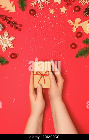 Frau mit Pastell Maniküre halten Handwerk Geschenkbox auf rotem Hintergrund mit weihnachtsdekoration und Schnee. Urlaubskonzept. Flach liegend, Kopierplatz Stockfoto