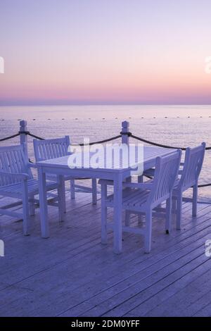 Sonnenaufgang, Sonnenuntergang Lila und Orange Lichter spiegeln sich auf dem leicht nassen weißen Holztisch und Stuhl am Pier in Antalya Türkei. Stockfoto