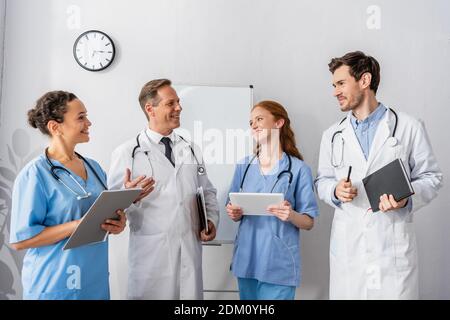 Glückliche multikulturelle Ärzte und Krankenschwestern, die einander anguckend Zusammen stehend mit Flipchart und Wanduhr auf dem Hintergrund Stockfoto