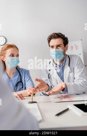 Arzt in medizinischer Maske im Gespräch mit Kollegen, während sie sitzen Arbeitsplatz mit Papieren und Gehirn anatomischen Modell auf verschwommenem Vordergrund Stockfoto