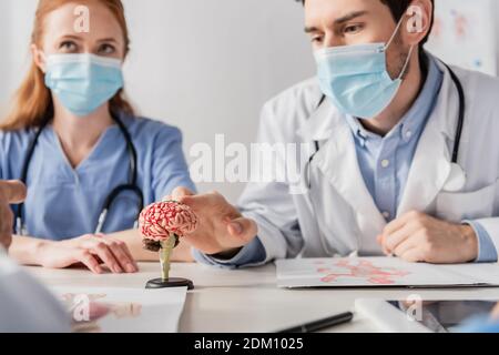 Arzt in medizinische Maske berühren Gehirn anatomischen Modell während des Sitzens Am Arbeitsplatz in der Nähe von Kollegen auf verschwommenem Vordergrund Stockfoto