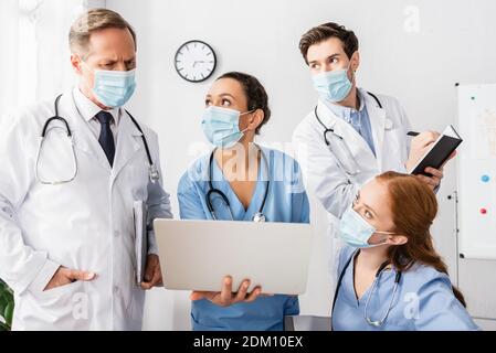 Arzt mit der Hand in der Tasche und dem Laptop in den Händen afroamerikanische Krankenschwester, während sie in der Nähe von Kollegen im Krankenhaus stand Stockfoto