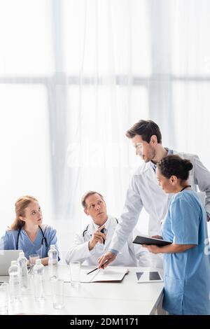 Multiethnische Krankenhauspersonal arbeitet mit Notebook, Geräten und Papieren in der Nähe von Wasserflaschen in der Klinik Stockfoto