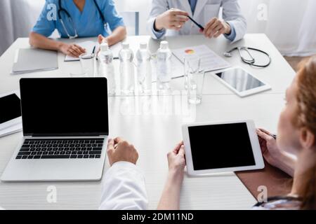Geräte mit leerem Bildschirm, Wasser und Papier auf dem Tisch in der Nähe des Krankenhauspersonals auf unscharfem Hintergrund Stockfoto
