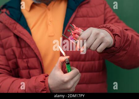 Mann in Rot aufgebockt Beleuchtung mehrere Feuerwerkskörper in seiner Hand mit Gasfeuerzeug. Guy immer bereit für das neue Jahr Spaß mit Feuerwerk oder pyrotechnische Produkte Stockfoto