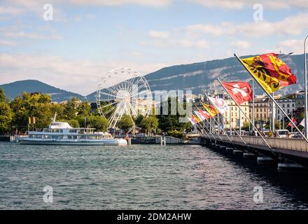 Genfer Flaggen und Schweizer Flaggen fliegen auf einer Brücke auf dem Genfer See mit einem Ausflugsboot, das am Fuße des Riesenrads festgemacht ist. Stockfoto