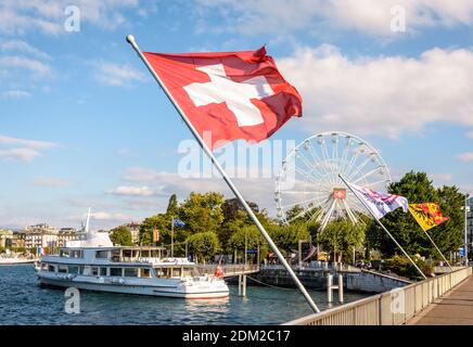 Die Schweizer Flagge fliegt im Wind auf dem Genfersee mit einem Ausflugsboot, das am Fuß des Riesenrads festgemacht ist. Stockfoto