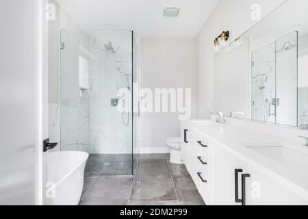 Ein modernes, weißes Luxusbad mit schwarzen Armaturen und Chromarmaturen. In der Ecke befindet sich eine große Dusche mit Glaswänden und Marmorfliesen. Stockfoto