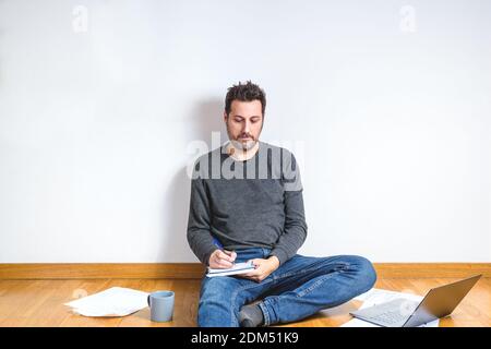 kaukasischer junger Mann mit hellem Bart auf dem Boden sitzend Von einer Wohnung und Schreiben auf einem Papier-Notizbuch Stockfoto