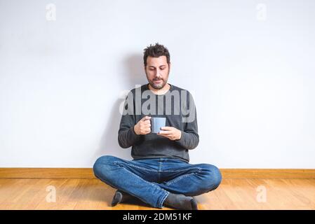 Junger Kaukasier, der auf dem Holzboden eines leeren Mannes sitzt Zimmer mit Kaffeetasse in den Händen Stockfoto