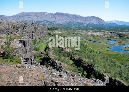 Blick über das Rifttal im Thingvellir Nationalpark, Island. Zerklüftete Felsen und Klippen neben flachen grünen Wiesen und ein Fluss fließt durch. Stockfoto