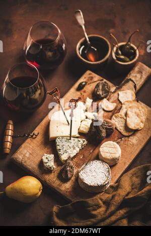 Auswahl an Snacks und Vorspeisen für Rotwein-Konzept. Verschiedene Käsesorten, Cracker, trockenes und frisches Obst und Gläser Rotwein auf rustikalem Holzbrett auf rostbraunem Hintergrund Stockfoto